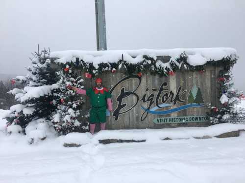 A snowy scene featuring a festive sign for Bigfork, with holiday decorations and a person dressed as an elf.