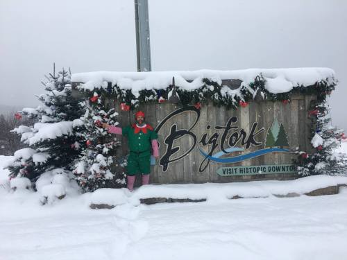 A snowy scene featuring a festive sign for Bigfork, with holiday decorations and a person dressed as an elf.