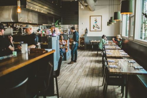 A modern restaurant interior with wooden floors, tables, and patrons dining, while staff work in the background.