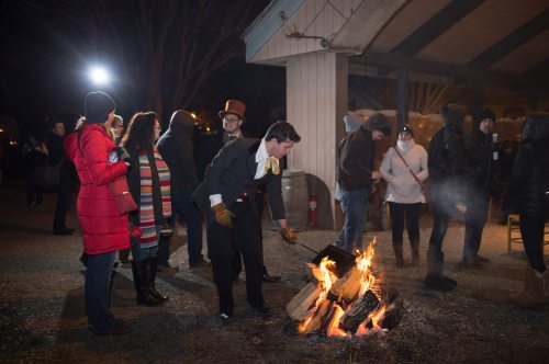 A group of people gathers around a fire at night, with one person in formal attire tending to the flames.