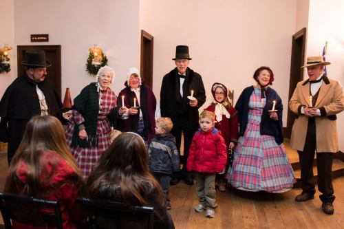 A group of people in historical costumes singing with candles, while children watch in a festive indoor setting.
