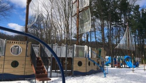 A playground resembling a ship, with slides and climbing structures, surrounded by snow and trees under a blue sky.