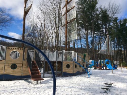 A playground designed like a ship, featuring slides and climbing structures, surrounded by snow and trees.