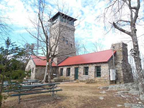 A stone building with a tall tower and red roof, surrounded by trees and a picnic area on a cloudy day.