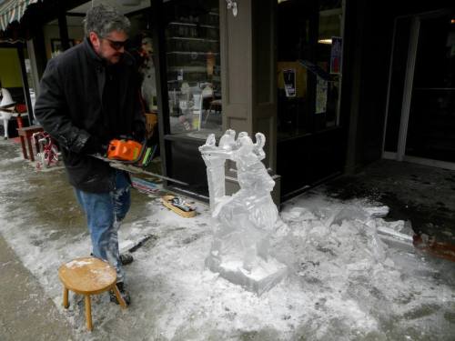 A man uses a chainsaw to carve an ice sculpture outside a shop, with ice shavings scattered on the ground.
