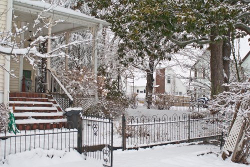 A snowy scene featuring a house with a porch, a fenced yard, and trees, creating a winter landscape.