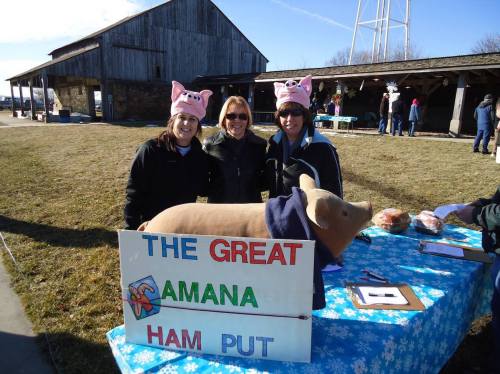 Three women wearing pig hats pose with a sign for "The Great Amana Ham Put" next to a pig statue outdoors.