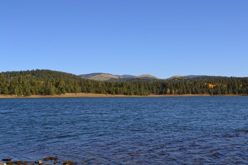 A serene lake surrounded by lush green forests and distant mountains under a clear blue sky.