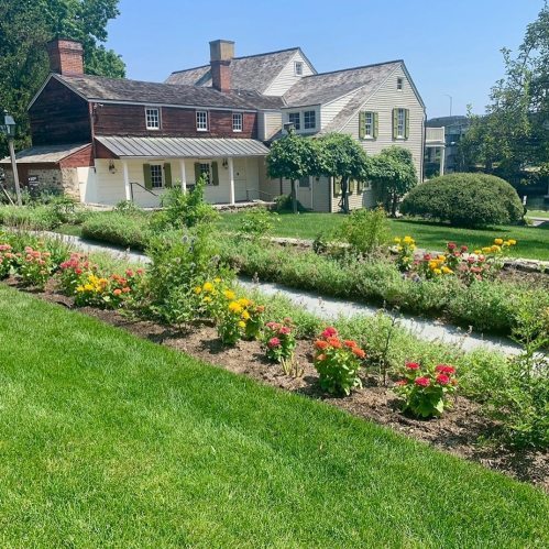 A sunny garden with colorful flowers in front of two historic houses and lush green grass.