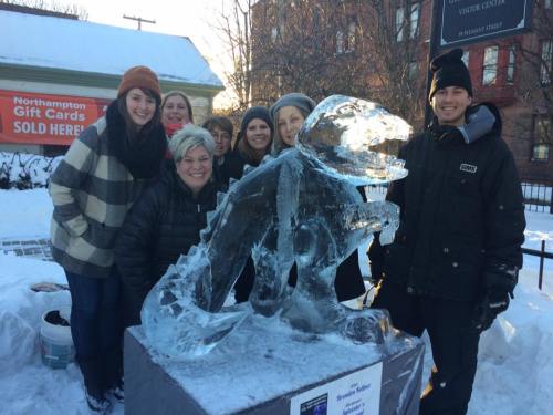 A group of people poses with a large ice sculpture of a dinosaur in a snowy outdoor setting.