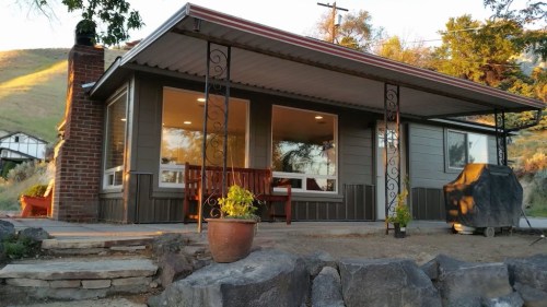 A cozy house with a porch, surrounded by hills, featuring a bench and a grill in the yard.
