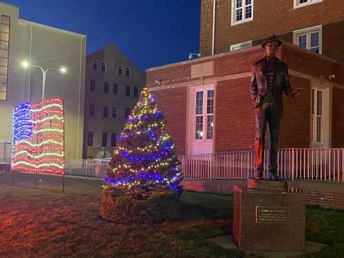 A decorated Christmas tree and a lighted American flag display beside a statue of a man in a cowboy hat at night.