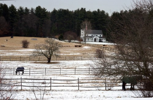 A snowy landscape with a farmhouse, trees, and a fenced pasture, surrounded by evergreen trees in the background.