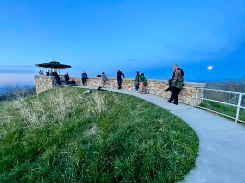 People gather on a scenic overlook at dusk, with a moon visible in the sky and grass surrounding the pathway.