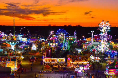 A vibrant carnival scene at sunset, filled with colorful lights, rides, and attractions against a glowing orange sky.