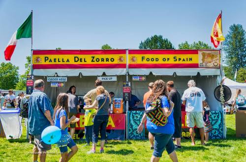 A vibrant food stall named "Paella Del Zorro" at an outdoor event, with people enjoying the sunny day.