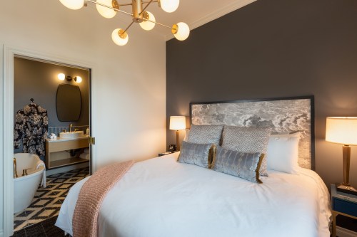 A stylish bedroom with a large bed, modern lighting, and an adjoining bathroom visible in the background.