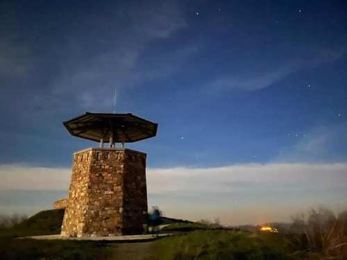 A stone observation tower under a starry night sky, with a faint glow from distant lights on the horizon.