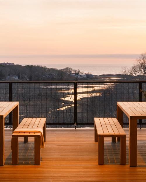 A wooden deck with tables and benches overlooks a serene sunset over the ocean and a winding river.