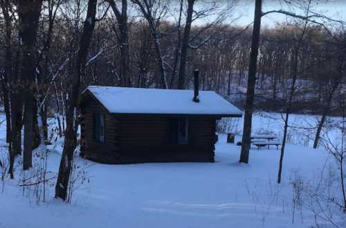 A small log cabin covered in snow, surrounded by trees in a winter landscape.