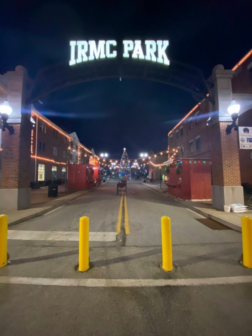 Night view of IRMC Park entrance, decorated with lights and a Christmas tree, with yellow bollards lining the street.