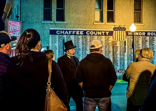 A group of people gathers at night, listening to a speaker in a top hat near a wall displaying names in Chaffee County.