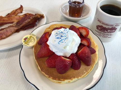 A plate of pancakes topped with strawberries and whipped cream, served with bacon, syrup, and a cup of coffee.