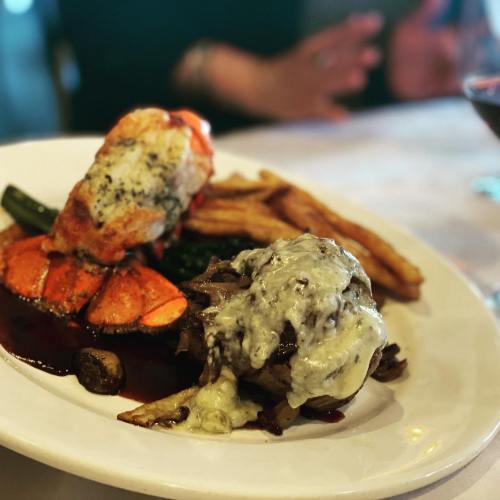 A plate of gourmet food featuring grilled lobster, vegetables, fries, and a meat dish topped with melted cheese.