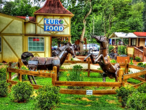 Metal horse sculptures in a fenced area outside a Euro Food shop, surrounded by greenery and a "Do Not Enter" sign.