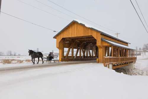 A horse-drawn sleigh passes under a wooden covered bridge in a snowy landscape.