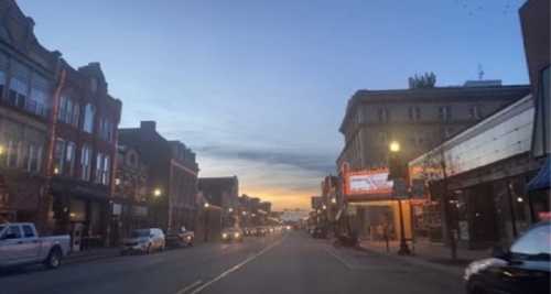 A quiet street at dusk, lined with buildings and parked cars, with a colorful sunset in the background.