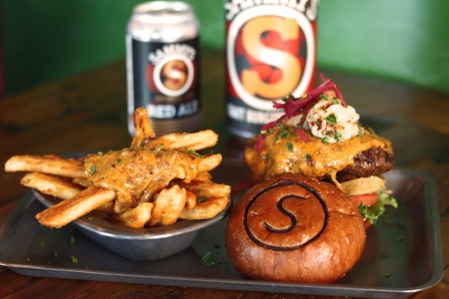 A plate with a cheesy burger topped with onions, served with fries and two cans of soda in the background.