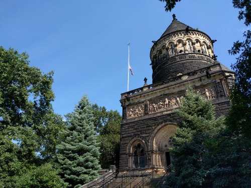 A historic stone building with a dome, surrounded by trees and a clear blue sky. An American flag flies at half-mast.