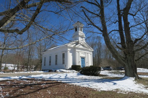 A white church with a steeple surrounded by snow and bare trees on a clear blue sky day.