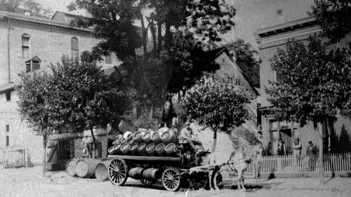 A historic black-and-white photo of a horse-drawn cart loaded with barrels in front of buildings and trees.