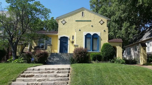 A bright yellow house with blue accents, surrounded by greenery and a stone pathway leading to the entrance.