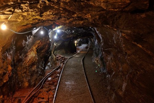 A dimly lit underground mine tunnel with rocky walls and a gravel path, featuring overhead lights and metal tracks.