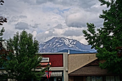 A snow-capped mountain rises behind buildings, framed by green trees under a cloudy sky.