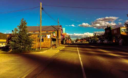 A quiet street in a small town, lined with wooden buildings and a clear blue sky with scattered clouds.