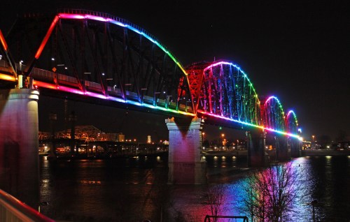 A brightly lit bridge at night, illuminated with colorful lights reflecting on the water below.