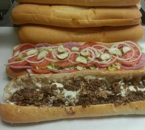 A close-up of two sub sandwiches: one with meats, veggies, and condiments, and another filled with shredded beef and onions.