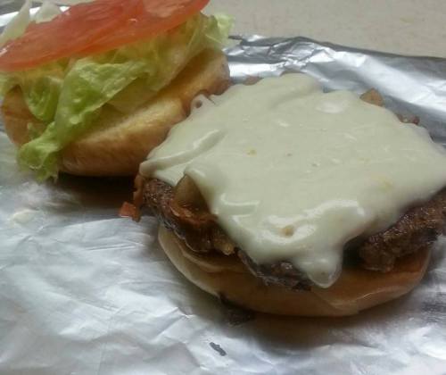 A sandwich with a bottom bun topped with melted cheese, sautéed onions, lettuce, and a slice of tomato on the side.