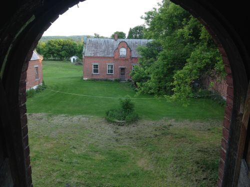 View through an archway of a grassy yard with a red brick building and trees in the background.