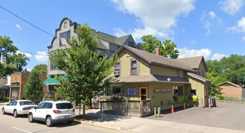A charming two-story building with a green exterior and purple accents, surrounded by trees and parked cars on a sunny day.