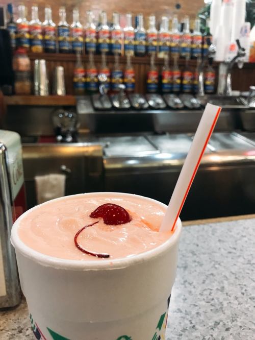 A close-up of a pink drink with a cherry on top and a straw, with colorful bottles in the background.
