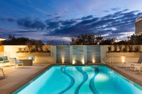 A serene pool area at dusk, featuring illuminated water, lounge chairs, and a backdrop of clouds and greenery.
