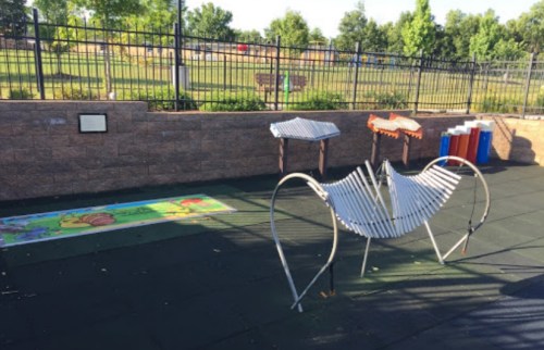 A playground area featuring musical instruments, a colorful mat, and a fenced grassy space in the background.