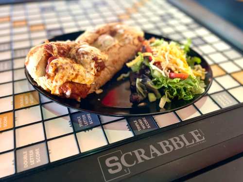 A plate with a cheesy sandwich and a fresh salad, set on a Scrabble game board.