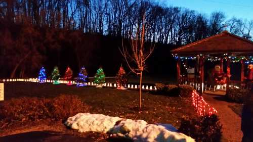 A festive scene with colorful Christmas lights, decorated trees, and a gazebo at dusk, surrounded by snow.