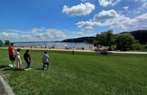 A family walks along a grassy path near a beach with people enjoying the water and a blue sky with fluffy clouds.